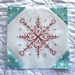 May include: Square white fabric block with a red embroidered snowflake design. The snowflake has heart-shaped details. The corners are accented with teal fabric triangles with white and green polka dots. The block is on a white quilted background.