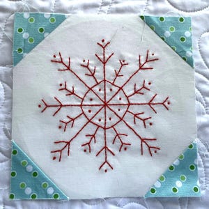 May include: A square fabric block with a hand-stitched red snowflake design on a white background. The corners are accented with triangles of blue fabric with green polka dots. This item is suitable for quilting or other craft projects.