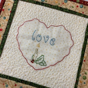 May include: A quilted square with a heart-shaped centerpiece. The heart is white with the word "love" embroidered in blue, and a candle and holly design. The border is green and patterned with red and gold accents.