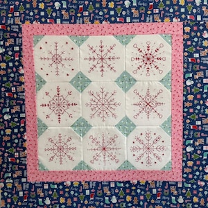 Puede incluir: Un edredón cuadrado hecho a mano con nueve bloques octogonales blancos, cada uno bordado con un diseño único de copo de nieve rojo. Los bloques están colocados sobre un fondo azul claro y enmarcados por un borde rosa y una tela azul oscuro estampada con motivos navideños.
