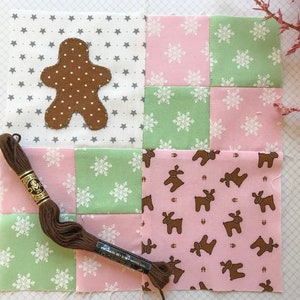 May include: A patchwork square with a brown gingerbread man applique, surrounded by fabric squares in pink, green, and gray. The fabrics feature star, snowflake, and reindeer patterns. A spool of brown embroidery floss rests on the fabric.