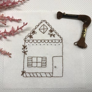 May include: A white fabric square featuring a brown embroidered house design. The house has a diamond window, a door, and floral details. A spool of brown embroidery thread is placed nearby, along with pink floral sprigs.