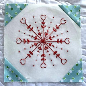 May include: A square fabric block with a red embroidered snowflake design. The snowflake features heart-shaped details and is centered on a white background. The corners are accented with triangles of blue fabric with green polka dots. This item is perfect for quilting.