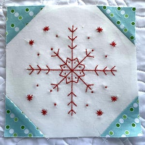 以下が含まれることがあります： 赤い刺繍の雪の結晶のデザインが施された白い中心部を持つ正方形の布ブロック。角は、白と緑のドット模様のターコイズ生地でアクセントが付けられています。このブロックは、より大きなキルトまたはクラフトプロジェクトの一部である可能性があります。
