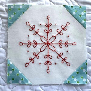 May include: A square fabric block with a white background and a red embroidered snowflake design. The corners are accented with triangles of blue fabric with white and green polka dots. This item is suitable for quilting or crafting.