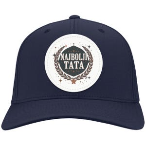 May include: Navy blue baseball cap with a circular patch. The patch features the text "NAJBOLJI TATA" in white, surrounded by a brown laurel wreath and stars. The cap has a curved brim and a structured crown.