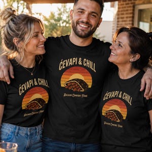 Peut inclure: Un t-shirt noir arborant un motif de coucher de soleil aux tons orange, rouge et jaune derrière un aliment. Le texte "ĆEVAPI & CHILL" est imprimé en arc au-dessus du soleil, avec "BALKAN COMFORT FOOD" écrit en plus petit en dessous.