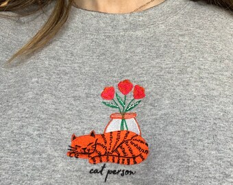 Puede incluir: Una sudadera gris con un diseño bordado de un gato naranja durmiendo junto a un jarrón de flores rojas. El texto "cat person" está bordado debajo del gato.