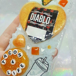 May include: A white phone case with a gold heart-shaped design featuring the text "DIABLO" and a taco. The case also has a round, gold circle with the text "TACOS 4 LIFE" and a black and white illustration of a cup with a straw.