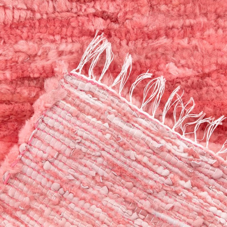 May include: Close-up of a coral-colored rug with a textured, plush surface. The rug's edge reveals a woven construction with white fringe detailing. The color palette is a soft, warm coral, creating a cozy aesthetic.