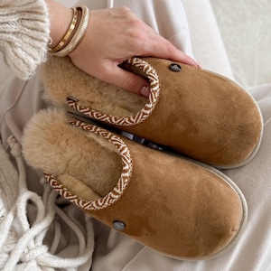 May include: A pair of brown suede slippers with a cream-colored fur lining and a decorative trim. The slippers have a round toe and a rubber sole. A small, round, silver-colored button is on the side of each slipper. The slippers are designed for indoor use.