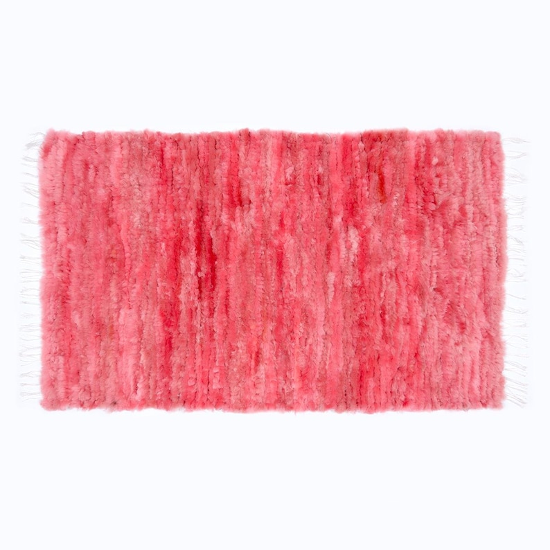 May include: A rectangular, plush rug in shades of pink. The rug has a textured surface with vertical lines and white fringe on the short ends. The rug appears soft and inviting.