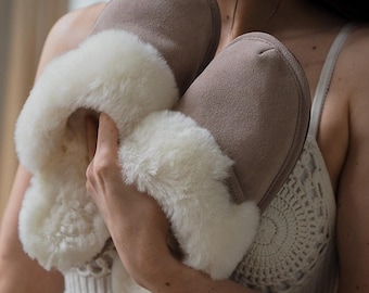 Può includere: Un paio di pantofole color tortora con una morbida fodera in pelliccia sintetica color crema che si estende sul bordo superiore. L'esterno ha una finitura liscia e opaca, mentre l'interno è completamente foderato in materiale bianco spesso.