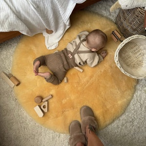 May include: A baby wearing a beige sweater and brown pants is playing on a round, yellow sheepskin rug. The baby is surrounded by wooden toys and there are two pairs of brown slippers in the background.