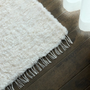 May include: A cream-colored, rectangular rug with a shaggy texture and fringe detail. The rug is made of a soft, plush material and is placed on a dark wood floor. The fringe is a lighter shade of white.