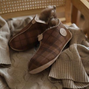 May include: A pair of brown plaid slippers with a fluffy lining. The slippers are sitting on a beige knitted blanket.