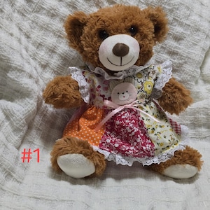 Peut inclure: Un ours en peluche marron portant une robe en patchwork avec des pois orange, des motifs floraux rouges et des fleurs jaunes. La robe présente une bordure en dentelle blanche, un nœud en ruban rose et un petit écusson en forme de chat. Le texte #1 est visible.