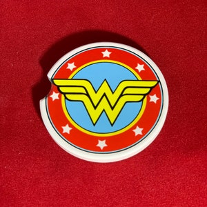 May include: A white ceramic coaster with a red, yellow, and blue Wonder Woman logo. The logo features a yellow W with wings and a blue circle with white stars around the edge.