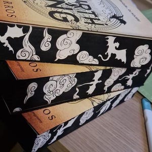 Puede incluir: Una pila de libros de fantasía con lomos negros que presentan ilustraciones de dragones y nubes blancas. Las portadas de los libros tienen un fondo beige con el título y el nombre del autor. Los libros están apilados en ángulo.