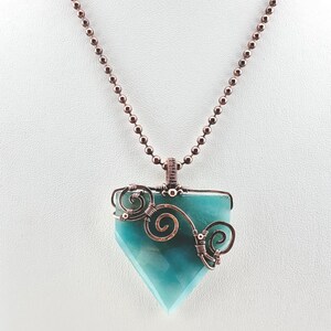 Può includere: Una collana con pendente con una pietra a forma di diamante azzurro chiaro. La pietra è avvolta in filo color rame con volute decorative. La catena della collana è composta da piccole perline color rame.
