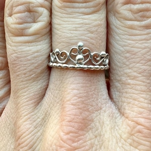 May include: A silver-colored ring in the shape of a crown. The ring features a beaded base and a delicate filigree design with small, round accents. The crown ring is worn on a finger.