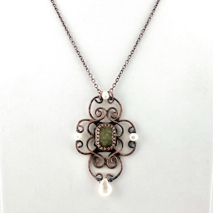 Op de afbeelding: Een koperkleurige hangerketting met een bloemmotief, een centrale groene steen en witte parelaccenten. De hanger heeft een open ontwerp met wervelende patronen en een delicate ketting.