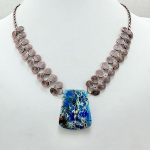 May include: A handcrafted necklace featuring a large, rectangular blue and white stone pendant. The pendant is suspended from a copper-toned chain with spiral design accents. The necklace is displayed on a white mannequin.