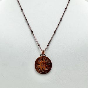 May include: A copper-colored pendant necklace. The round pendant features an embossed geometric design with a central starburst pattern. The necklace chain is a matching copper color with small bead accents.