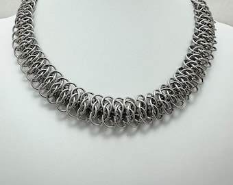 May include: A silver-toned chainmail necklace constructed from numerous interlocking metal rings. The necklace features a dense, uniform weave pattern that creates a thick, rounded collar shape around the neck area.