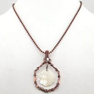 May include: A copper-toned necklace featuring a pendant with a white seashell design. The pendant is suspended within a wire-wrapped frame, and the necklace has a beaded chain. The seashell pendant is a pearly white color.
