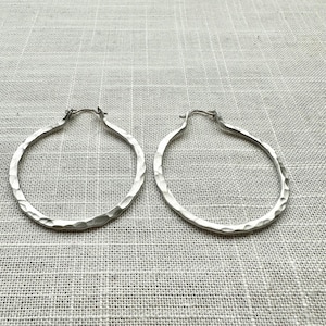 May include: A pair of silver hoop earrings with a hammered texture. The earrings are circular and have a simple, elegant design. The background is a light gray fabric.