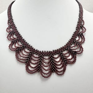 Puede incluir: Un collar de cuentas de color marrón rojizo con un diseño de conchas. El collar está hecho de pequeñas cuentas redondas que están enhebradas juntas en un patrón que crea una serie de conchas en cascada.