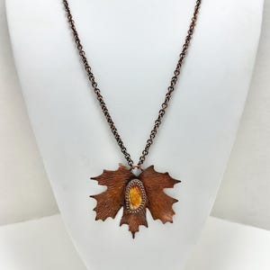 May include: A copper necklace with a maple leaf pendant featuring a brown and yellow stone in the center.