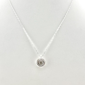 Könnte beinhalten: Eine silberne Halskette mit einem kreisförmigen Anhänger. Der Anhänger besteht aus ineinander verschlungenen Silberringen, die ein strukturiertes, geknotetes Design bilden. Die feine Kette hängt vor einem weißen Hintergrund.