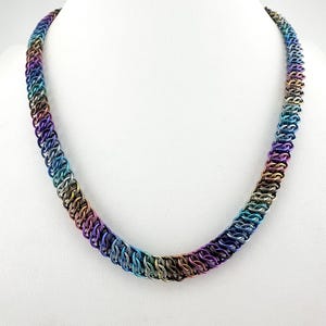 May include: A colorful chainmail necklace displayed on a white mannequin. The necklace is made of small, interlocking metal rings in a rainbow of colors, including purple, blue, green, and gold. The chainmail is woven into a thick, rounded shape.