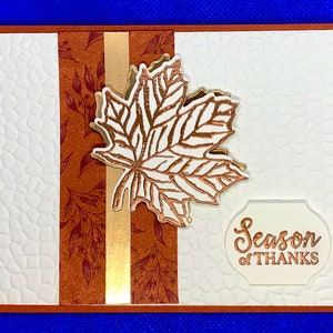 May include: A white card with a brown and gold patterned border. A gold foil leaf is on the card with the text "Season of THANKS" in gold foil on a white tag.