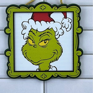 May include: A framed Christmas ornament featuring the Grinch's face. The Grinch is green with a red Santa hat and white trim. The frame is green with a decorative edge, and a gold cord is attached for hanging.