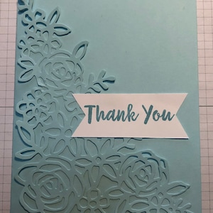 May include: A light blue card with a white banner that says "Thank You". The card has a decorative floral design in the corner.