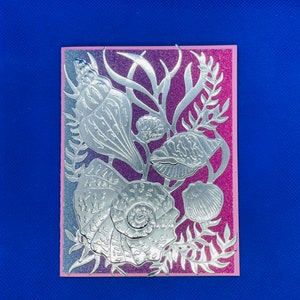 Puede incluir: Una tarjeta plateada con un fondo degradado rosa y morado. La tarjeta presenta un diseño de corte plateado de conchas marinas y algas.