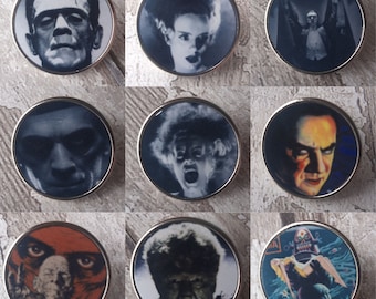 May include: Nine round, silver-colored pins with black and white images of classic horror movie characters. The characters include Frankenstein's monster, the Creature from the Black Lagoon, Dracula, and the Wolfman.