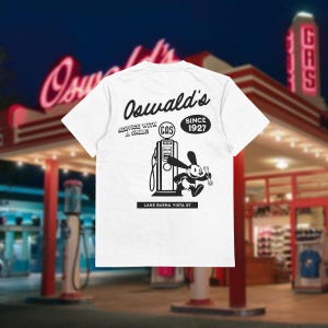 Può includere: T-shirt bianca con grafica in bianco e nero di un coniglio dei cartoni animati con una chiave inglese vicino a una pompa di benzina. La maglietta presenta il testo "Oswald's", "Service With A Smile", "Since 1927" e "Lake Buena Vista St."