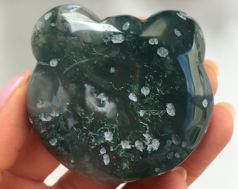 Puede incluir: Piedra de ágata verde musgo en forma de corazón con inclusiones blancas. La piedra se sostiene en una mano.