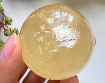Op de afbeelding: Een grote, gladde, gele kristallen bol met iriserende regenboogflitsen. De bol wordt in een hand gehouden.
