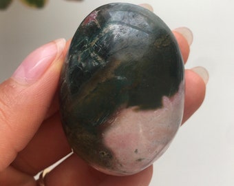 Può includere: Una pietra preziosa verde e rosa levigata e liscia con un motivo a spirale. La pietra ha una forma ovale e ha un aspetto naturale e organico.