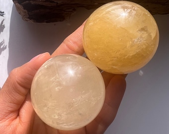 May include: Two crystal spheres, one is a light yellow color and the other is a translucent white. The spheres are sitting on a hand.