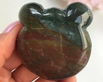 Puede incluir: Una piedra de jaspe pulida de color verde, marrón y rojo, tallada en forma de oso. La piedra tiene un acabado liso y brillante.