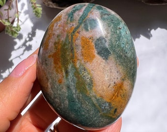 May include: A smooth, polished green, orange, and pink jasper palm stone. The stone has a swirling pattern of colours and is a natural, earthy gemstone.