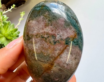 May include: A smooth, polished green, brown, and pink jasper palm stone. The stone has a swirling pattern of colours and is a natural, organic shape.