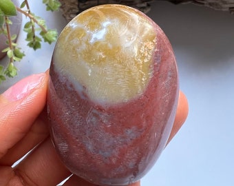 Può includere: Una pietra levigata di forma ovale, con una miscela di colori. La parte superiore è giallo crema, che sfuma in una fascia bianca, e poi in una tonalità rosso-marrone scuro. La pietra ha una superficie liscia e lucida, tenuta in una mano.