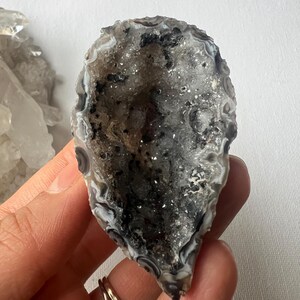 Puede incluir: Una geoda de ágata gris y blanca con un exterior con bandas blancas y negras. La geoda está abierta y revela un interior forrado de cristales.